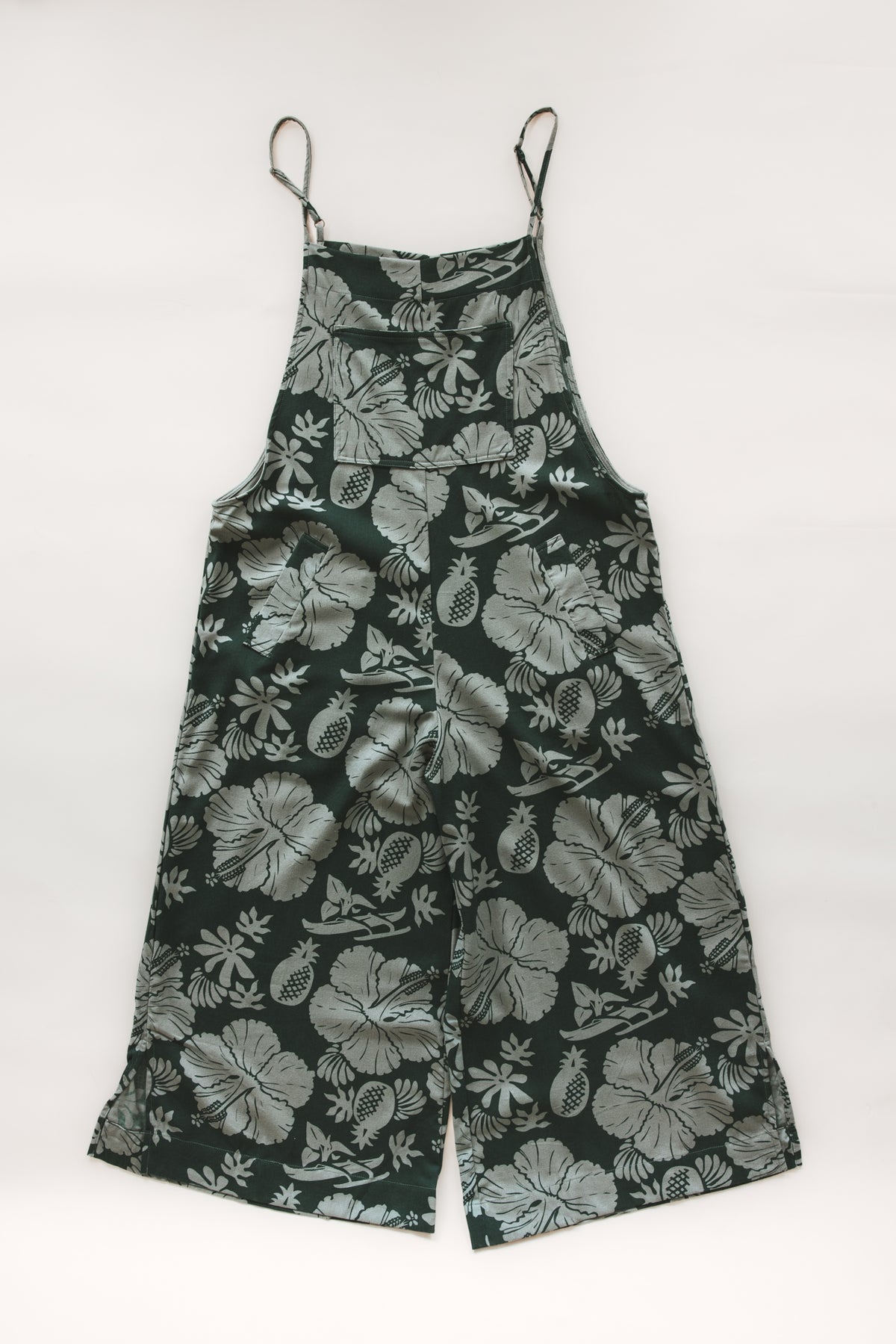 Wings Hawaii women's aloha print overalls in Hana Forest. Designed in Maui, made in Bali. Dark green tropical pattern with hibiscus florals, pineapples, and Hawaiian outrigger canoes. Adjustable straps, wide leg, relaxed fit, front and side pockets. Small batch, sustainable clothing for beachy, boho style. 