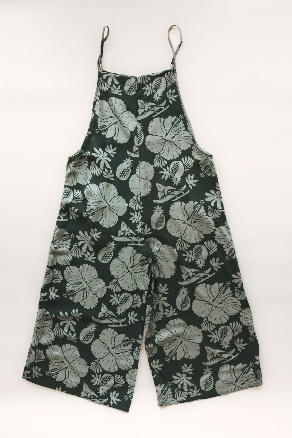 Wings Hawaii women's aloha print overalls in Hana Forest. Designed in Maui, made in Bali. Dark green tropical pattern with hibiscus florals, pineapples, and Hawaiian outrigger canoes. Adjustable straps, wide leg, relaxed fit, front and side pockets. Small batch, sustainable clothing for beachy, boho style. 