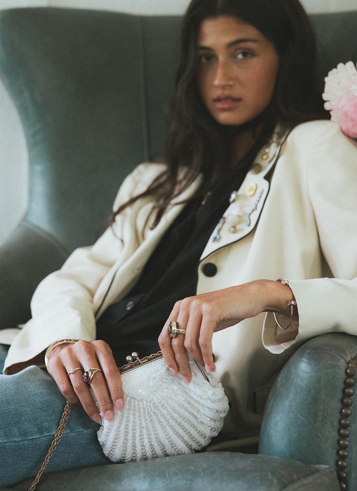 Model in a cream blazer holding a shell purse in a green armchair